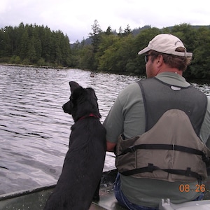 Puede incluir: Un hombre con un chaleco salvavidas y una gorra de béisbol se sienta en un bote con un perro negro. El perro está mirando el agua. El hombre está mirando al perro. El agua está tranquila y hay árboles en el fondo.