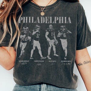 Puede incluir: Una camiseta de manga corta gris carbón con el texto PHILADELPHIA en una fuente desgastada sobre cuatro jugadores de fútbol americano en uniforme. Los nombres Saquon Barkley, Cooper DeJean, Jalen Hurts y DeVonta Smith aparecen impresos.