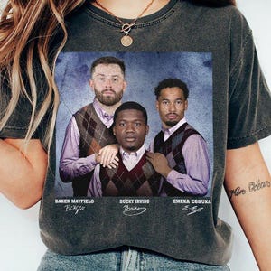 May include: A charcoal gray t-shirt featuring a graphic print of three men wearing light purple dress shirts and brown argyle sweater vests. The names Baker Mayfield, Bucky Irving, and Emeka Egbuka are printed below each person with their signatures.