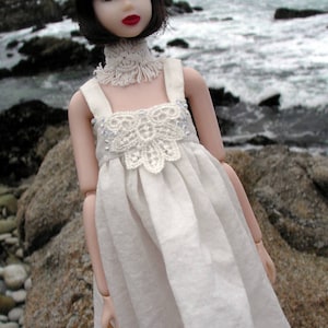 May include: A doll with black hair and red lipstick wearing a white dress with a crocheted bodice and collar. The doll is standing in front of a rocky shoreline. The text "PDF Digital Pattern" is visible in the upper right corner.
