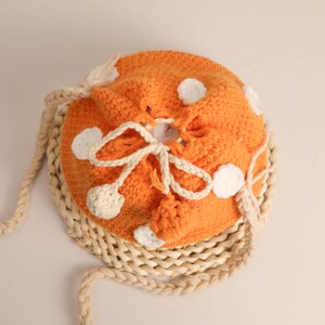 May include: An orange crocheted drawstring bag with white polka dots and a cream-colored drawstring closure. The bag has a cream-colored braided strap and is sitting on a woven mat. The bag is handmade.
