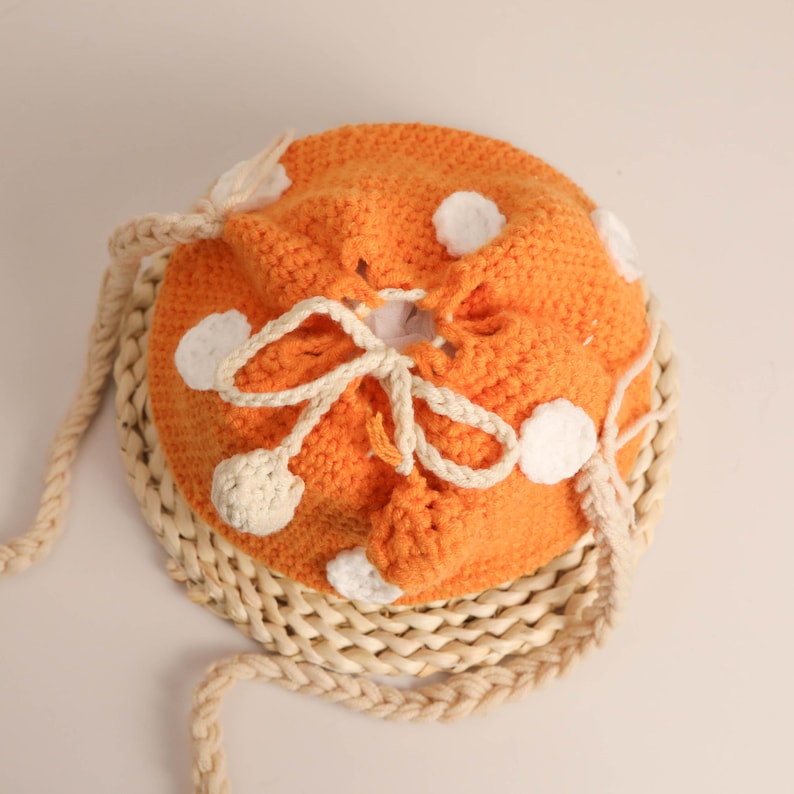 May include: An orange crocheted drawstring bag with white polka dots and a cream-colored drawstring closure. The bag has a cream-colored braided strap and is sitting on a woven mat. The bag is handmade.
