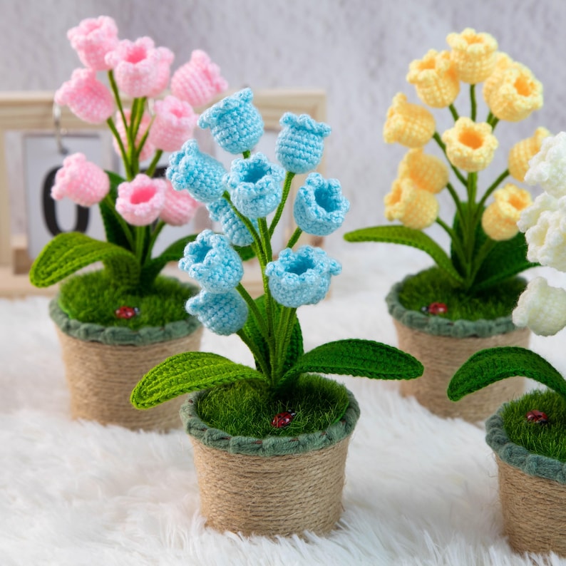 K&ouml;nnte beinhalten: Geh&auml;kelte Blumenarrangements in kleinen T&ouml;pfen. Die Blumen sind in Rosa, Blau und Gelb gehalten, mit gr&uuml;nen Bl&auml;ttern und Stielen. Die T&ouml;pfe sind mit Bindfaden umwickelt und mit gr&uuml;nem Moos gef&uuml;llt. Ein kleiner Marienk&auml;fer ist auf einigen T&ouml;pfen zu sehen.
