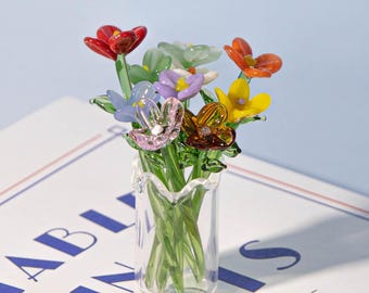May include: A miniature glass vase holding a vibrant bouquet of glass flowers. The flowers are in various colours, including red, orange, yellow, purple, blue, and green. The clear vase has a ruffled edge. The flowers are arranged in a natural, organic style.