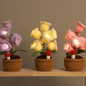 Puede incluir: Tres lámparas de flores de ganchillo en morado, amarillo y rosa, cada una con un pequeño hongo rojo y blanco. Las flores están en macetas tejidas marrones, y las lámparas están sobre una estantería de madera. Las lámparas están encendidas.