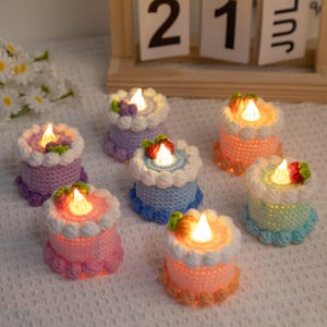 Könnte beinhalten: Eine Sammlung von sieben kleinen, gehäkelten LED-Lichtern in Kuchenform. Jeder Kuchen hat eine andere Pastellfarbe, darunter Rosa, Blau, Lila und Orange, mit weißem Zuckerguss und einer kleinen, leuchtenden LED-"Kerze" oben. Die Kuchen sind auf einer weißen Oberfläche angeordnet.