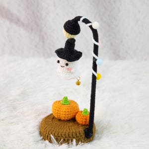 Può includere: Decorazione di Halloween all'uncinetto con un fantasma sorridente con un cappello da strega sotto un lampione nero. Due zucche arancioni sono posizionate su una base marrone all'uncinetto. Il lampione è decorato con perline colorate e una piccola campana.
