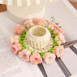 Puede incluir: Un posavasos hecho a mano, de ganchillo blanco roto, con un portavasos central, rodeado de flores de ganchillo color melocotón y hojas verdes. El posavasos está colocado sobre un libro.