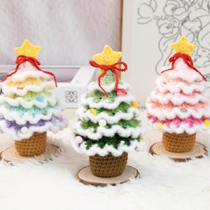 May include: Three handmade crochet Christmas trees in rainbow, green, and pink hues. Each tree features a yellow star topper, red bow, and sits on a wooden base. The trees are crafted with detailed stitching and colorful yarn.