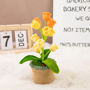 K&ouml;nnte beinhalten: Ein handgefertigtes H&auml;kelblumenarrangement mit gelben und orangen Tulpen in einem kleinen Topf, der mit Bindfaden umwickelt ist. Der Topf hat gr&uuml;ne Bl&auml;tter und einen kleinen Marienk&auml;fer. Ein Holzkalender zeigt das Datum 7. Dezember an.