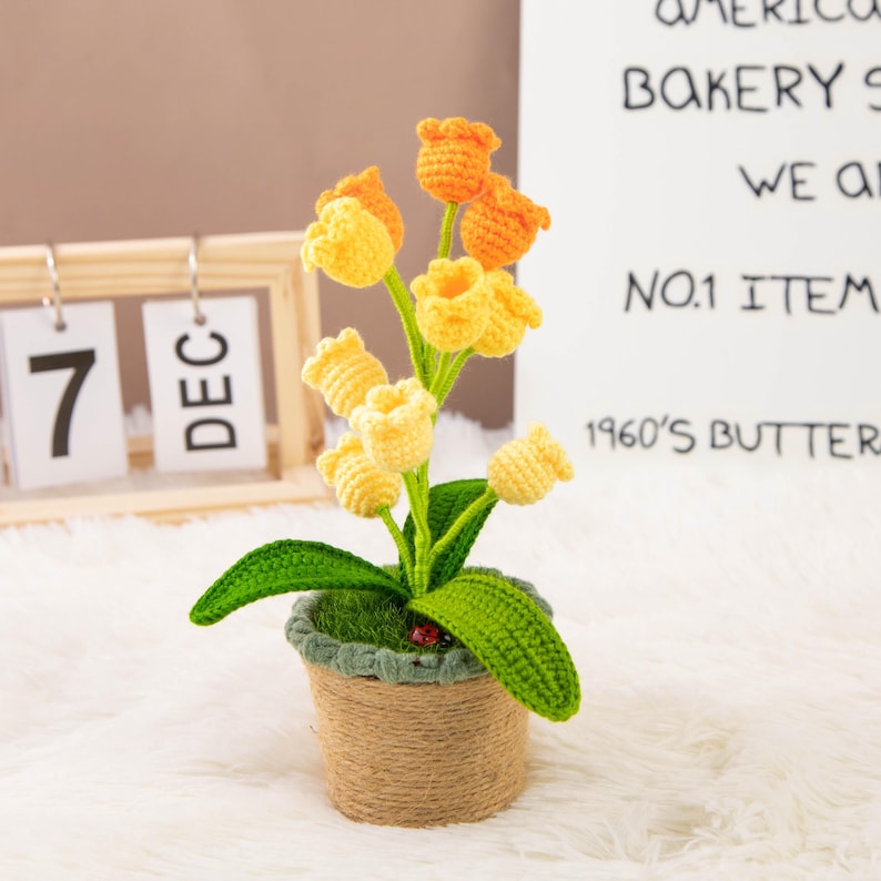 K&ouml;nnte beinhalten: Ein handgefertigtes H&auml;kelblumenarrangement mit gelben und orangen Tulpen in einem kleinen Topf, der mit Bindfaden umwickelt ist. Der Topf hat gr&uuml;ne Bl&auml;tter und einen kleinen Marienk&auml;fer. Ein Holzkalender zeigt das Datum 7. Dezember an.