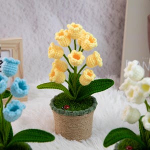K&ouml;nnte beinhalten: Ein handgefertigtes H&auml;kelblumenarrangement mit gelben glockenf&ouml;rmigen Bl&uuml;ten, gr&uuml;nen Bl&auml;ttern und einem kleinen roten Marienk&auml;fer in einem mit Jute umwickelten Topf. Die Anordnung befindet sich vor einem weichen, wei&szlig;en Hintergrund, mit anderen H&auml;kelblumen im Hintergrund.