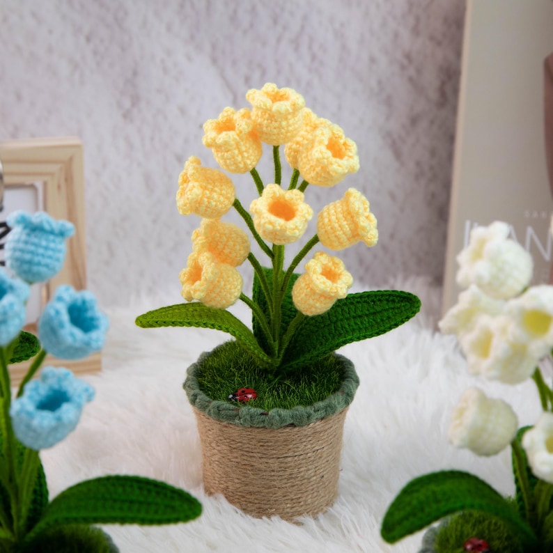K&ouml;nnte beinhalten: Ein handgefertigtes H&auml;kelblumenarrangement mit gelben glockenf&ouml;rmigen Bl&uuml;ten, gr&uuml;nen Bl&auml;ttern und einem kleinen roten Marienk&auml;fer in einem mit Jute umwickelten Topf. Die Anordnung befindet sich vor einem weichen, wei&szlig;en Hintergrund, mit anderen H&auml;kelblumen im Hintergrund.