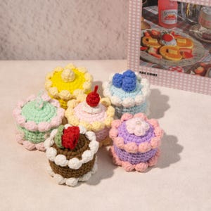 Puede incluir: Una colección de seis mini pasteles de ganchillo en varios colores, incluyendo amarillo, verde, rosa y marrón. Cada pastel presenta elementos decorativos como cerezas, fresas y arándanos. Los pasteles están dispuestos en grupo, mostrando sus detalles hechos a mano.
