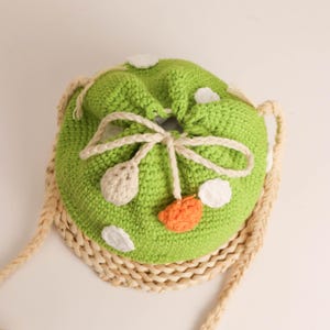 May include: A green crochet drawstring bag with white and orange accents. The bag features a woven base and a beige braided strap. The bag is decorated with white and orange crochet details, and a beige bow.