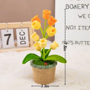 K&ouml;nnte beinhalten: Ein handgefertigtes H&auml;kelblumenarrangement in einem gewebten Topf. Das Arrangement zeigt gelbe und orangefarbene tulpenartige Bl&uuml;ten, gr&uuml;ne Bl&auml;tter und einen kleinen Marienk&auml;fer. Der Topf ist etwa 8,4 cm breit und das Arrangement ist 19,8 cm hoch.