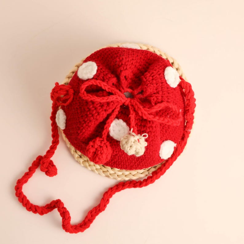 May include: A red crocheted mushroom-shaped bag with white polka dots and a braided red strap. The bag has a drawstring closure and a beige pom-pom accent. The bag sits on a woven, circular base.