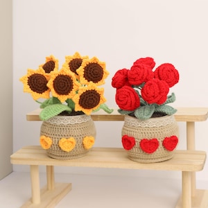 May include: Two crochet flower pots, one with sunflowers and the other with red roses. The pots are tan with lace trim and heart accents. The sunflowers have yellow petals and brown centers. The roses are a vibrant red.