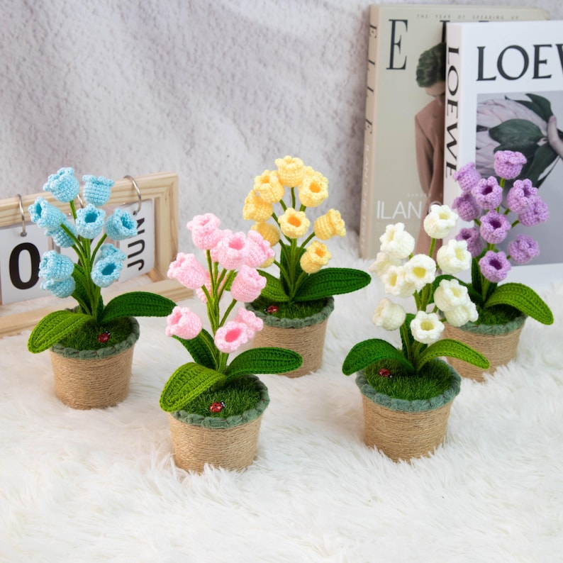 K&ouml;nnte beinhalten: F&uuml;nf geh&auml;kelte Blumenarrangements in kleinen T&ouml;pfen. Die Blumen sind in verschiedenen Farben wie Blau, Rosa, Gelb, Wei&szlig; und Lila gehalten. Jeder Topf ist mit Bindfaden umwickelt und mit gr&uuml;nem Moos gef&uuml;llt. Im Hintergrund ist ein kleiner Holzkalender zu sehen.