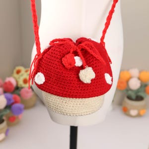 May include: A red and white crocheted mushroom-shaped bag with a drawstring closure and a long red strap. The bag has white polka dots and a beige base. The bag is displayed on a mannequin.