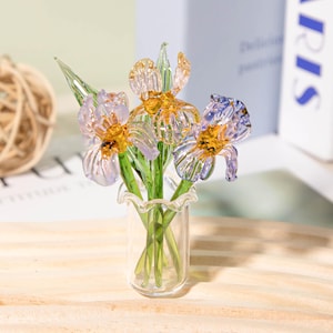 Può includere: Una composizione floreale in vetro con tre fiori di iris in un vaso trasparente. Gli iris hanno petali viola e gialli con steli e foglie verdi. Il vaso ha un bordo a balze.