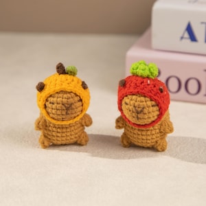 May include: Two small, handmade crochet figures. One figure wears a yellow hat with brown and green accents, the other wears a red hat with green accents. Both figures are brown and have a bear-like appearance. The figures are displayed on a light-colored surface.