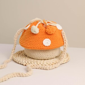 May include: An orange and beige crochet mushroom-shaped bag with a braided strap. The mushroom cap has white polka dots and small pom-poms. The bag sits on a woven beige base.