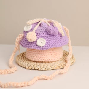 May include: A handmade crochet mushroom-shaped bag in shades of purple and peach. The bag features white and peach-colored polka dots and a braided strap. The bag is sitting on a woven mat.