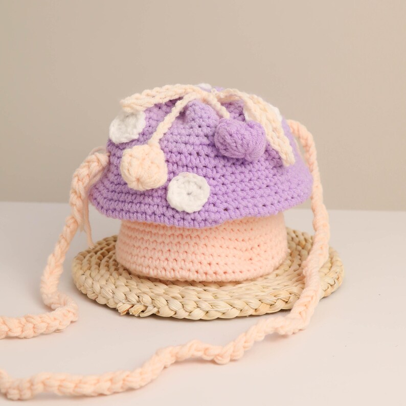 May include: A handmade crochet mushroom-shaped bag in shades of purple and peach. The bag features white and peach-colored polka dots and a braided strap. The bag is sitting on a woven mat.