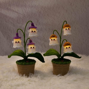 May include: Two crochet ghost flower lamps. Each lamp features a green stem with leaves, a pot wrapped in twine, and glowing ghost figures wearing purple or orange hats. The lamps are set on a fluffy white surface.