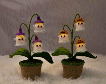 May include: Two crochet ghost flower lamps. Each lamp features a green stem with leaves, a pot wrapped in twine, and glowing ghost figures wearing purple or orange hats. The lamps are set on a fluffy white surface.