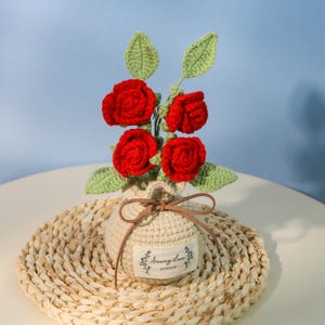 Könnte beinhalten: Ein handgefertigtes gehäkeltes Blumenarrangement mit vier roten Rosen und grünen Blättern. Die Blumen befinden sich in einer beige gehäkelten Vase mit einem braunen Band, die auf einer gewebten Matte steht. Das Etikett auf der Vase lautet "Sewing Lace Studio".