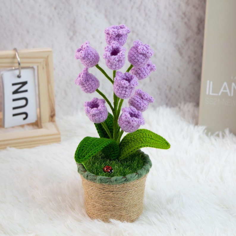 K&ouml;nnte beinhalten: Ein handgefertigtes H&auml;kelblumenarrangement mit lavendelfarbenen, glockenf&ouml;rmigen Bl&uuml;ten, gr&uuml;nen Bl&auml;ttern und Stielen. Die Blumen sind in einem kleinen, mit Bindfaden umwickelten Topf gepflanzt. Ein kleiner roter Marienk&auml;fer ist auf dem gr&uuml;nen Moos zu sehen.