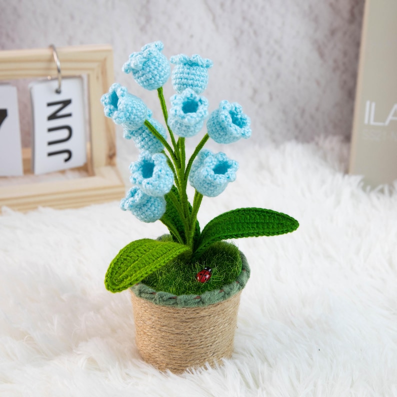 K&ouml;nnte beinhalten: Eine handgefertigte H&auml;kelblumenanordnung mit hellblauen, glockenf&ouml;rmigen Bl&uuml;ten, gr&uuml;nen Bl&auml;ttern und einem kleinen Marienk&auml;fer. Die Blumen befinden sich in einem braunen Topf, der mit Bindfaden umwickelt ist. Ein dekorativer Artikel.