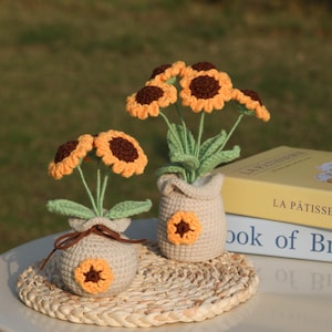 May include: Two crochet sunflower bouquets in beige pots with brown centers and yellow petals. The sunflowers are displayed on a woven mat. The pots have a sunflower applique. The background is blurred.