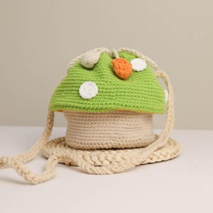 May include: A whimsical, handmade crochet mushroom-shaped bag. The bag features a light green cap with white polka dots and an orange accent. The stem is beige, and the bag has a matching strap. The bag sits on a woven, circular base.