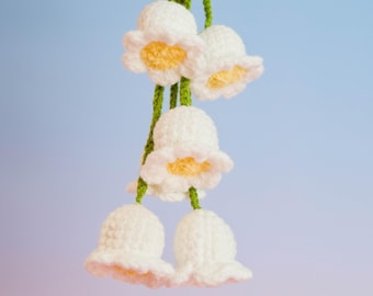 May include: A handmade crochet floral garland featuring white bell-shaped flowers with yellow centers. The flowers are strung together with green yarn, creating a delicate and charming decorative piece. The background is a gradient of blue and pink.