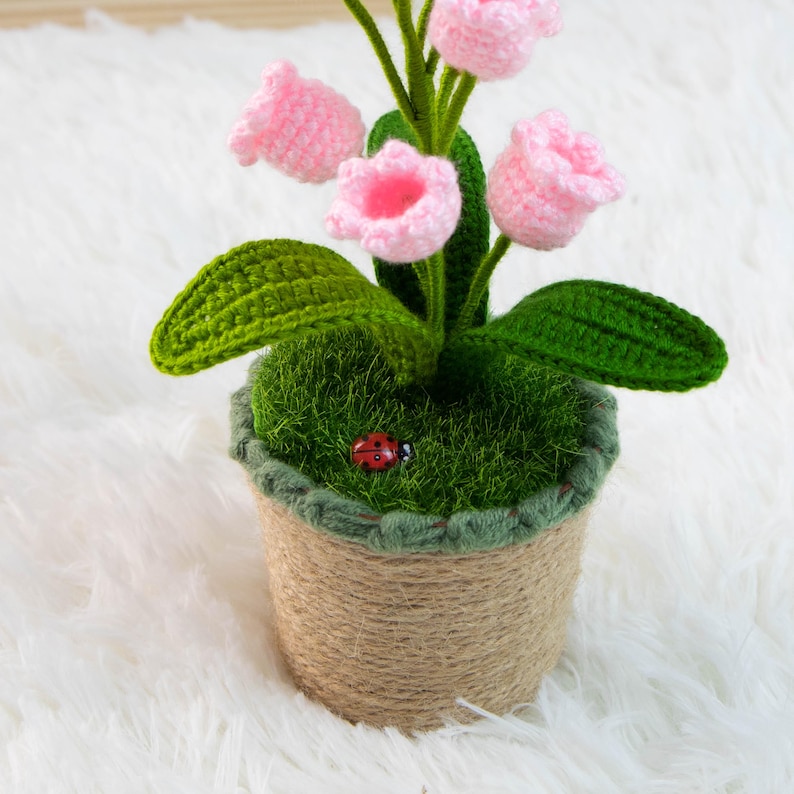 K&ouml;nnte beinhalten: Ein handgefertigtes H&auml;kelblumenarrangement in einem Topf. Das Arrangement zeigt rosa glockenf&ouml;rmige Bl&uuml;ten, gr&uuml;ne Bl&auml;tter und einen kleinen roten Marienk&auml;fer. Der Topf ist mit Bindfaden umwickelt und mit gr&uuml;nem Moos gef&uuml;llt.