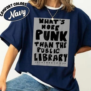 May include: Navy blue t-shirt with the text "WHAT'S MORE PUNK THAN THE PUBLIC LIBRARY" in a black, bold font on a gray rectangle. The shirt also has the words "COMFORT COLORS" and "Navy" in a navy blue oval.