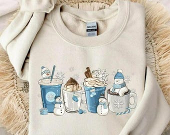 May include: Cream-colored sweatshirt featuring a winter-themed graphic. The design includes various beverages like coffee and hot chocolate, each adorned with snowmen and snowflakes. The sweatshirt has long sleeves and a crew neck.