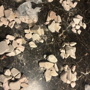 May include: A collection of broken pieces of light pink and white clay, scattered on a dark brown surface. The clay pieces are various sizes and shapes.