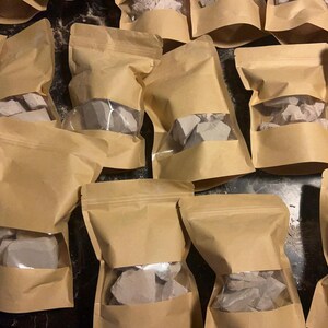 May include: Multiple brown paper bags filled with white clay chunks. The bags are sealed and have a clear window to see the contents.