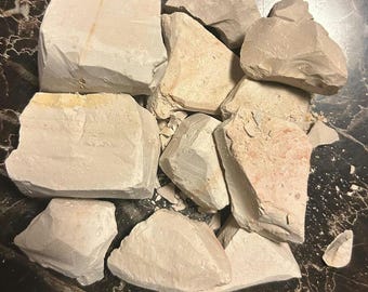 Puede incluir: Una colección de trozos irregulares y dentados de arcilla cruda en tonos beige claro y blanco roto. Las piezas varían en tamaño y forma, presentando texturas mate y calcáreas con sutiles manchas minerales de color rojo anaranjado.