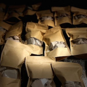 May include: A close-up of a pile of brown paper bags filled with small, grey, irregularly shaped rocks. The bags are sealed and have a clear window to see the contents.