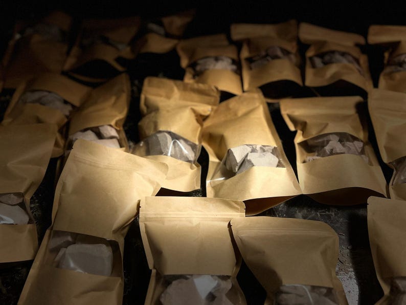 May include: A close-up of a pile of brown paper bags filled with small, grey, irregularly shaped rocks. The bags are sealed and have a clear window to see the contents.