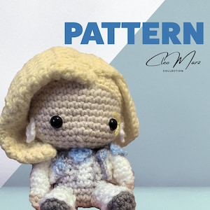 Könnte beinhalten: Eine gehäkelte Amigurumi-Puppe mit blonden Haaren, die ein weiß-blaues Outfit trägt. Die Puppe sitzt mit gekreuzten Beinen und hat ein süßes, cartoonartiges Gesicht.