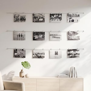 Peut inclure: Un mur d'exposition composé de trois câbles métalliques horizontaux fixés avec des pinces, supportant douze photographies en noir et blanc disposées en grille. Un buffet en bois clair avec une plante et des livres se trouve en dessous.