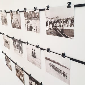 Peut inclure: Une présentation de photographies en noir et blanc fixées à des rubans noirs contre un mur blanc. Les photos représentent diverses scènes, notamment des personnes sur une plage et sur une place de ville. Les clips ont la forme de ciseaux.