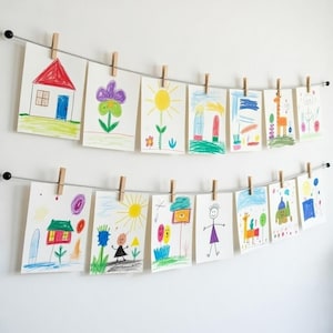 May include: Two horizontal metal wires mounted to a wall, each holding a row of white paper sheets with colorful crayon drawings. The drawings feature various subjects including houses, flowers, suns, animals, and stick figures, all secured by wooden clips.