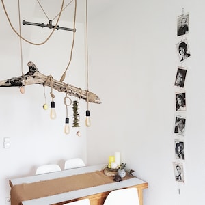 Op de afbeelding: Een rustieke, handgemaakte lamp hangt boven een eettafel. De lamp is gemaakt van een stuk drijfhout, touw en Edison-stijl gloeilampen. Een reeks zwart-wit portretten hangt aan de muur. De tafel heeft een bruine loper.